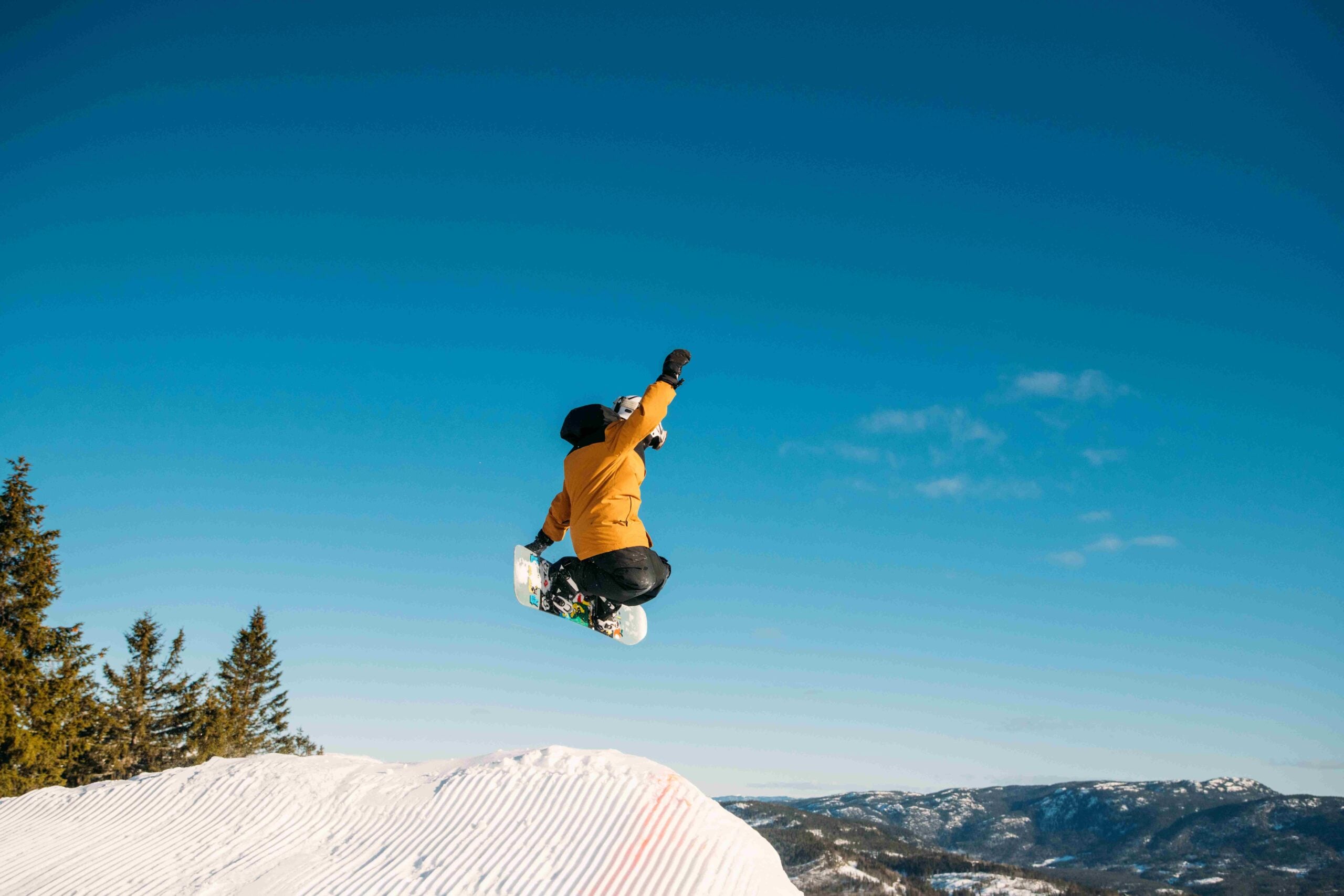 Ung snowboarder utfører et stort hopp over en snøbakke med blå himmel i bakgrunnen.